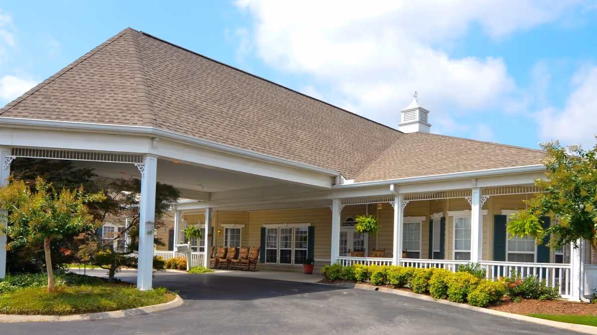 Exterior view of a single-story building with a large covered entrance supported by white columns. The building has a brown shingled roof, beige siding, white trim, and several windows with green shutters. There are rocking chairs on the porch, hanging plants, and landscaped bushes and trees around the entrance. The sky is partly cloudy.