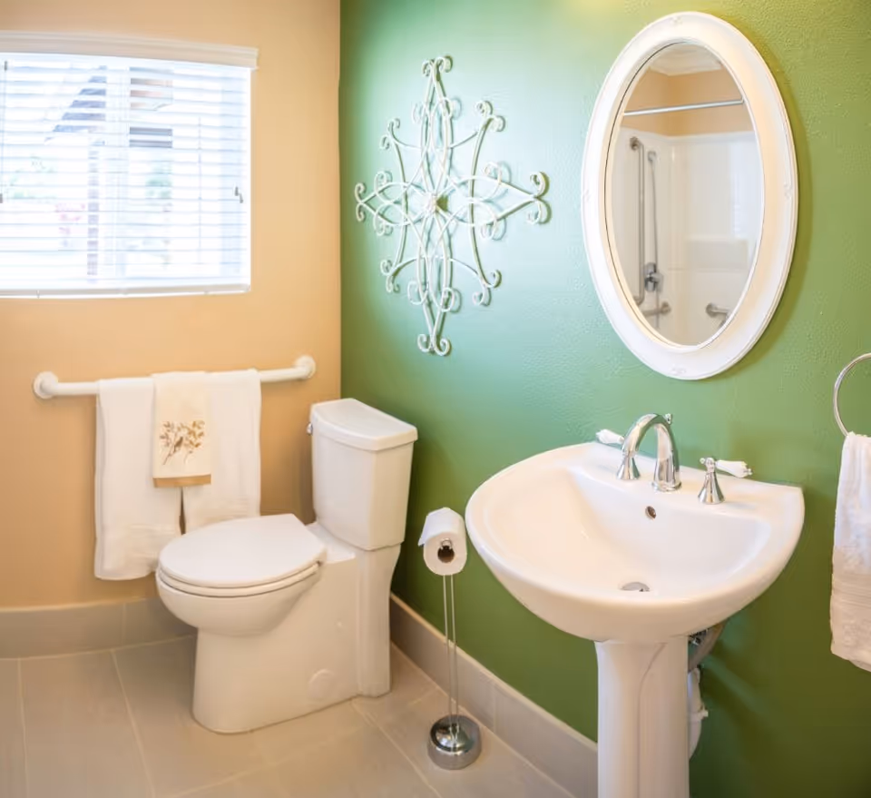 A clean bathroom with a white toilet and pedestal sink. The walls are painted green and beige, with a decorative metal wall art piece above the toilet. A round mirror hangs above the sink, and there are white towels on a towel rack and ring. A window with white blinds lets in natural light.