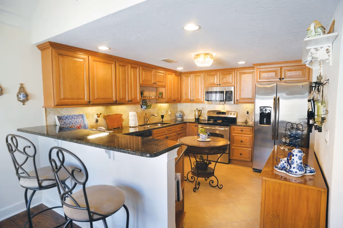 A well-lit kitchen with wooden cabinets, a black granite countertop breakfast bar with two metal bar stools, a round wooden table with decorative items, stainless steel appliances including a refrigerator, microwave, and stove, and various kitchen accessories and decor.