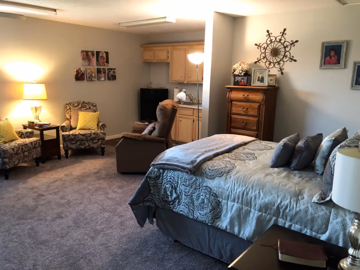 A cozy bedroom with a large bed covered in patterned bedding and multiple pillows. To the right of the bed is a nightstand with a lamp and a book. In the background, there is a wooden dresser with framed photos and decorative items on top. On the left side of the room, there are two upholstered armchairs with yellow accent pillows, a small side table with a lamp, and a brown recliner chair. The room has carpeted flooring and soft lighting, with a small kitchenette area featuring cabinets, a sink, and a mini refrigerator. Family photos and wall decor are displayed on the walls.