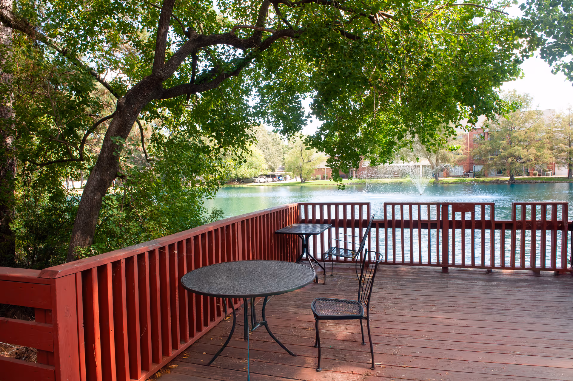 A red wooden deck with metal tables and chairs overlooking a lake with a fountain and leafy trees.