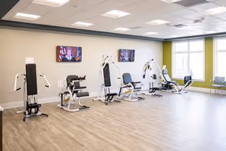 A spacious exercise room with light wood flooring and pale walls, equipped with multiple white and black strength training machines lined up against one wall. Two flat-screen TVs are mounted on the wall above the machines. Large windows on the adjacent wall let in natural light, and a few chairs are placed near the windows.