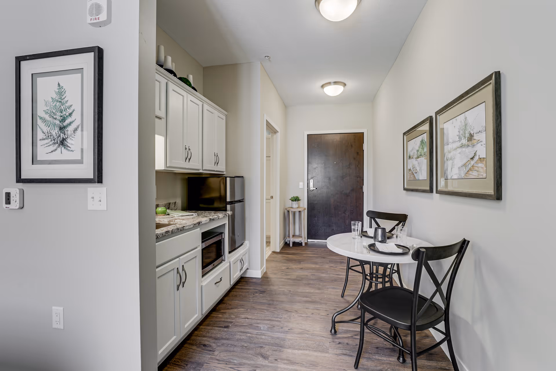 A small dining area with a round white table and two black chairs set against a light gray wall with two framed pictures. To the left, there is a compact kitchen area with white cabinets, a microwave, and a small refrigerator. The floor is wooden, and a dark wooden door is visible at the end of the hallway.