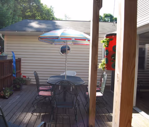 Outdoor wooden deck area with a black metal table and six matching chairs with red cushions. A colorful striped umbrella is open above the table. There are potted plants along the wooden railing and a red decorative hanging planter on a wooden post. The deck is adjacent to a beige siding building with a small octagonal window.