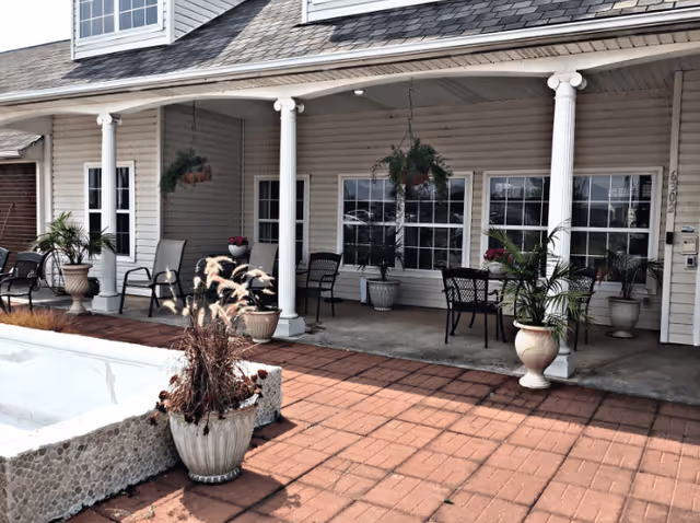 Outdoor patio area of The Chateau of Lawton with several chairs and potted plants arranged under a covered porch supported by white columns. The building has beige siding and multiple windows, with a brick-paved floor and a small empty stone planter in the foreground.