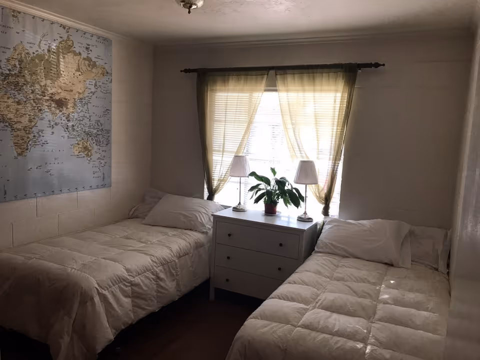 Small bedroom with two twin beds, a dresser with lamps and a potted plant between them and a world map on the wall.