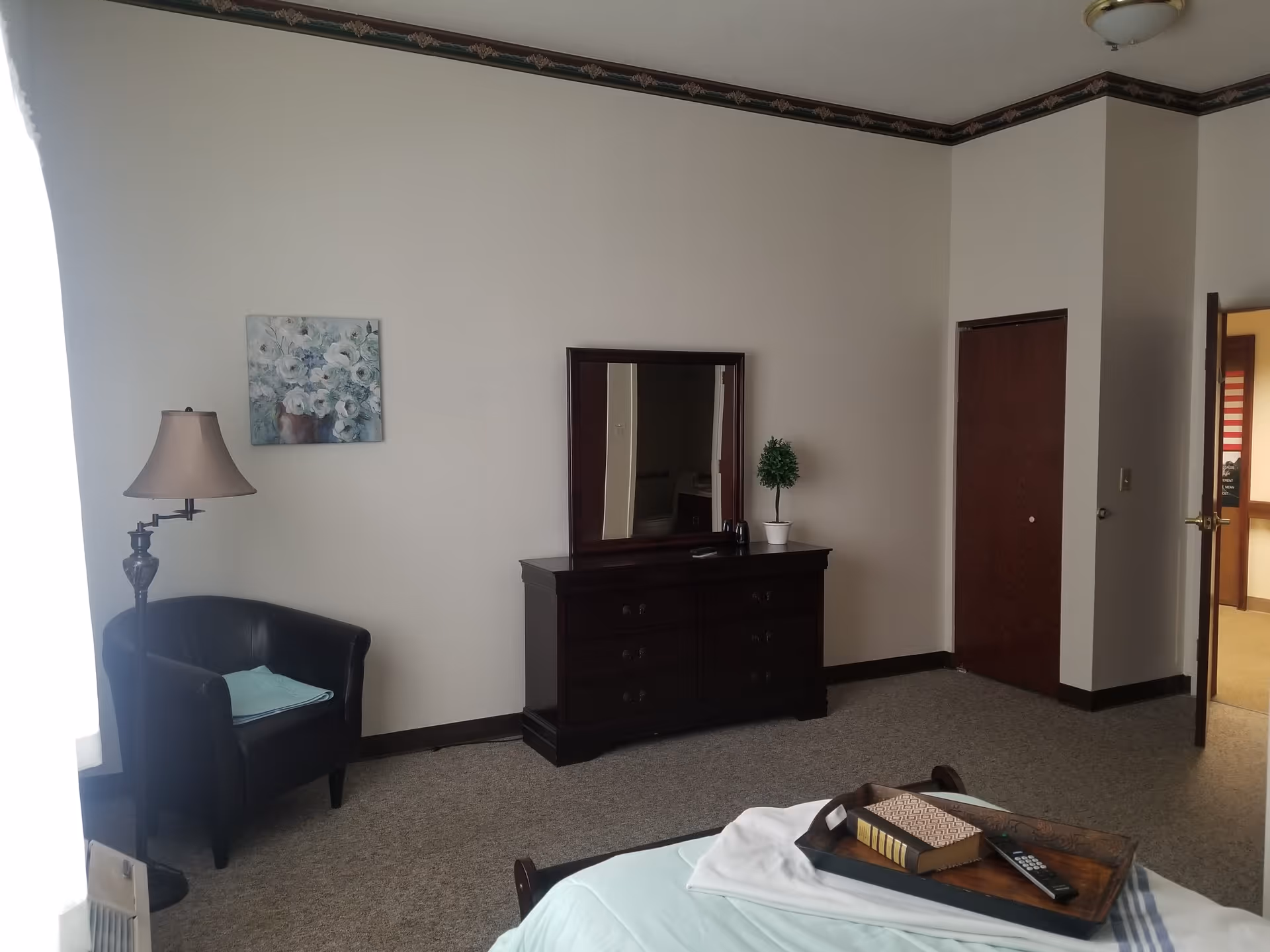 A senior living bedroom with a bed in the foreground holding a tray with a book and remote control. The room has a dark wooden dresser with a mirror, a small potted plant, a black armchair with a folded blanket, a floor lamp, and a floral painting on the wall. There is a door open to a hallway and a closed closet door.