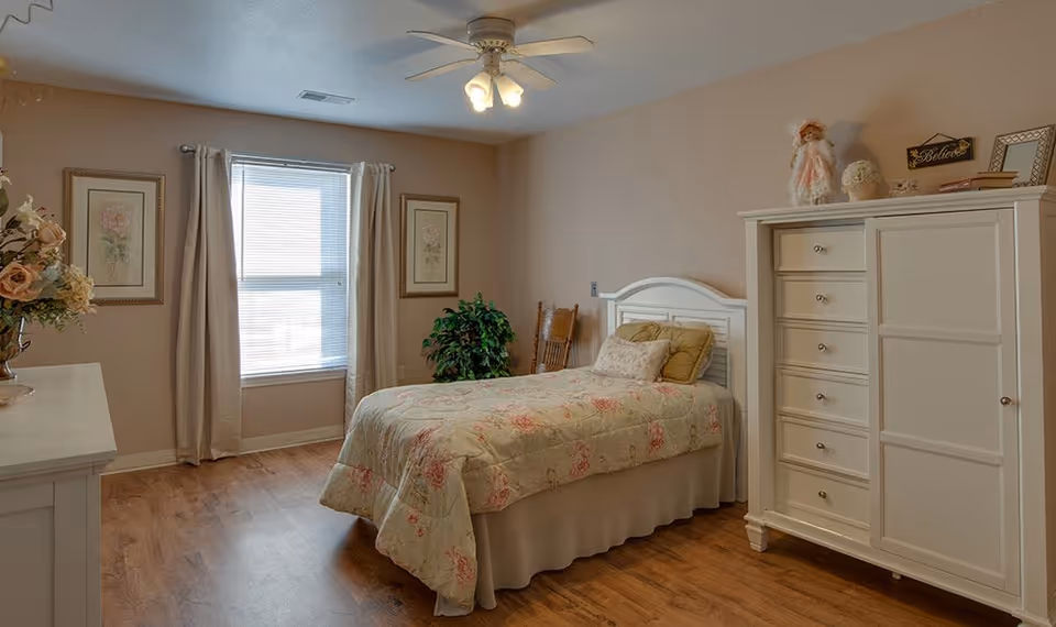 A cozy bedroom with a single bed covered in a floral quilt, a white dresser with decorative items on top, a wooden chair, two framed floral pictures on the wall, a window with beige curtains, and a ceiling fan with lights. The floor is wooden and the walls are painted beige.