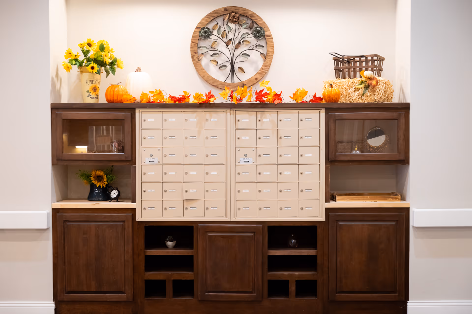 A wooden mail organizer with multiple small mail slots, decorated with autumn-themed items including pumpkins, fall leaves, and sunflowers. Above the mail slots is a round wall decoration featuring a metal tree with flowers. The organizer has cabinets and shelves on either side with additional decorative items.