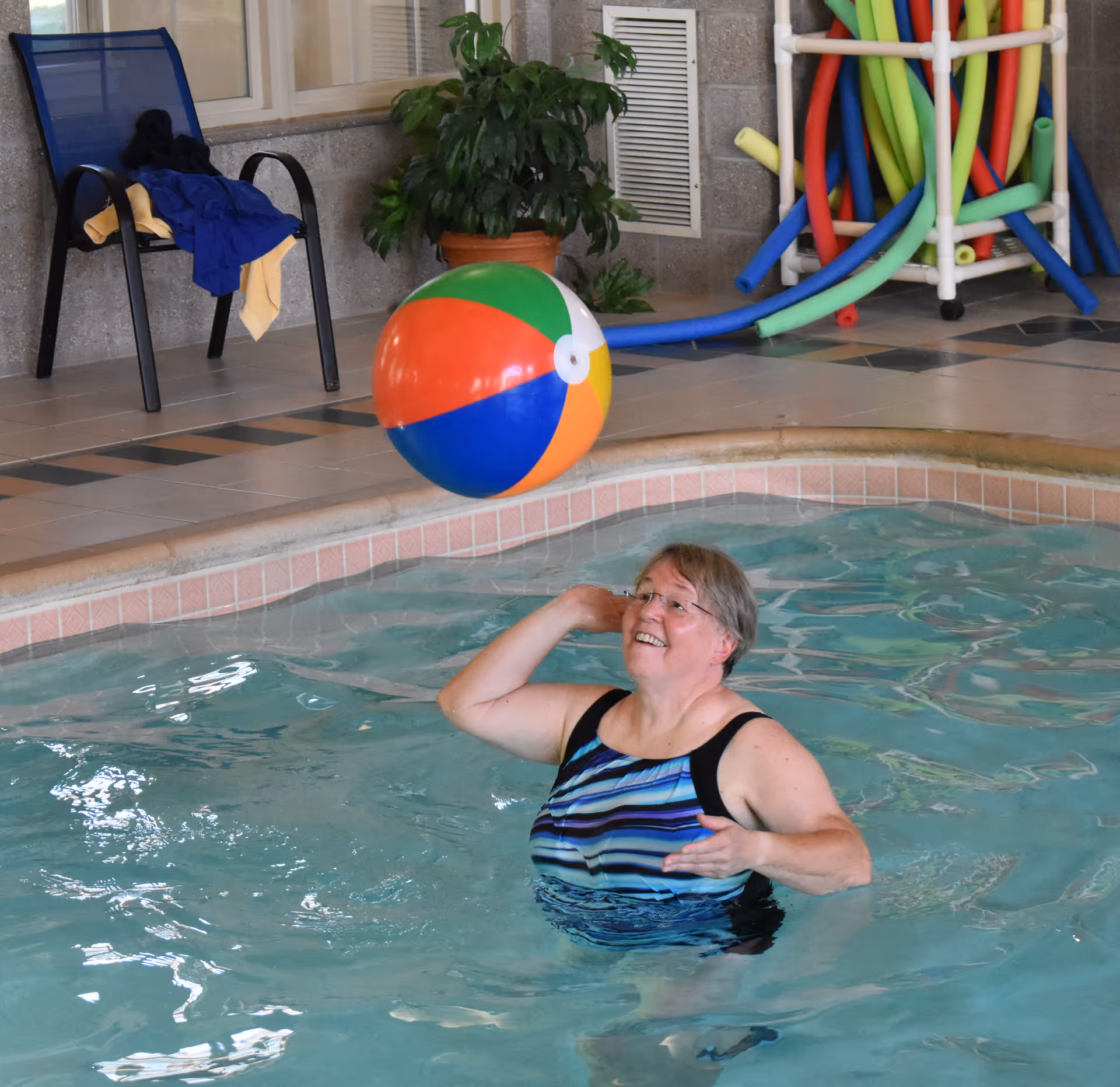 An older woman in a striped swimsuit is playing with a colorful beach ball in an indoor swimming pool. In the background, there is a blue chair with towels on it, a potted plant, and a rack holding colorful pool noodles.