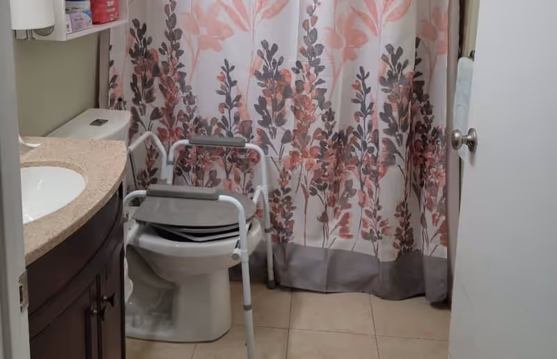 Bathroom with a toilet, sink vanity, floral shower curtain, and an over-toilet transfer/commode frame.