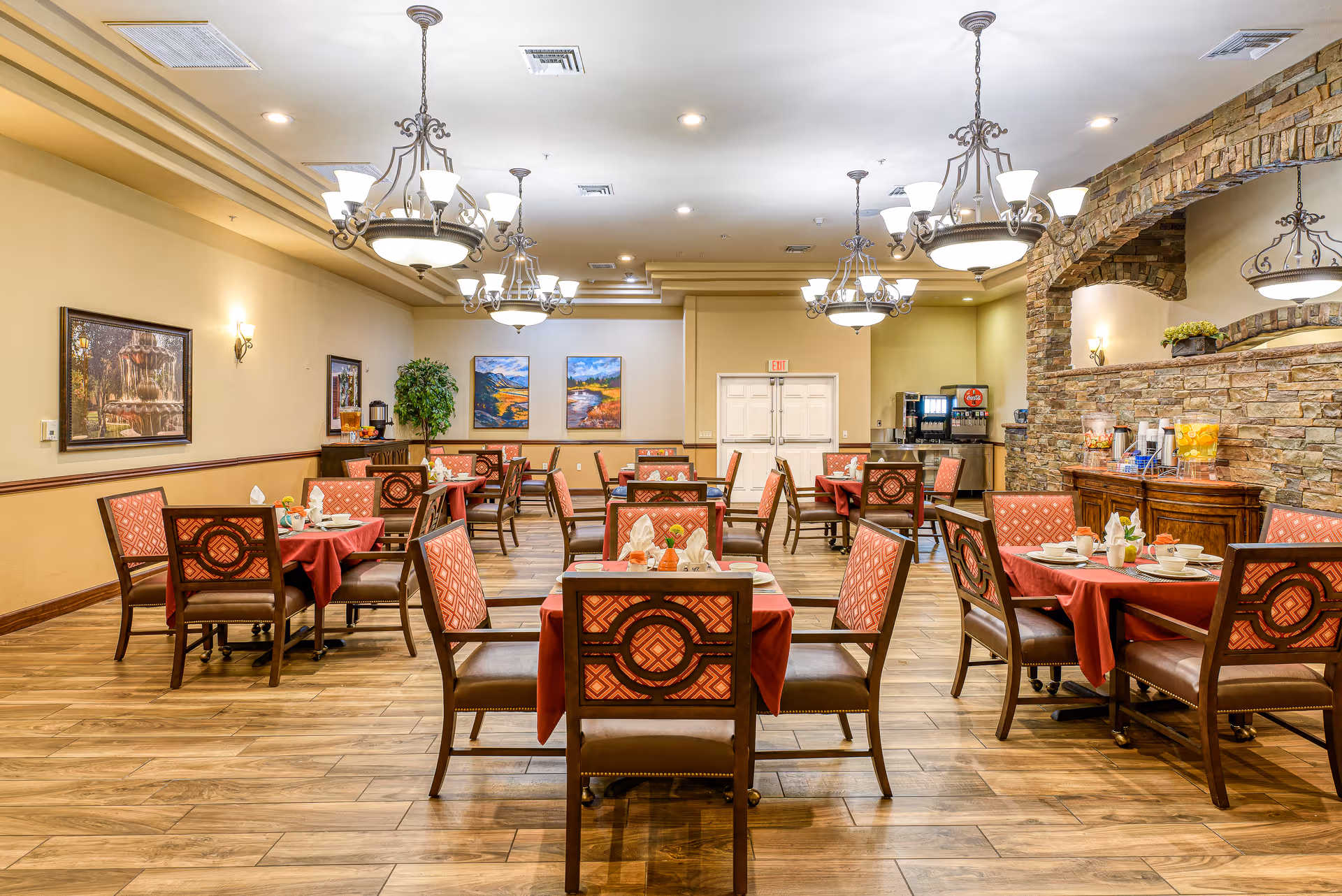 A spacious dining room with multiple tables covered with red tablecloths and set with white napkins, plates, and cups. The chairs have patterned upholstery and wooden frames. The room features wooden flooring, warm beige walls, decorative chandeliers, framed artwork, and a stone archway with a sideboard holding beverage dispensers and cups.
