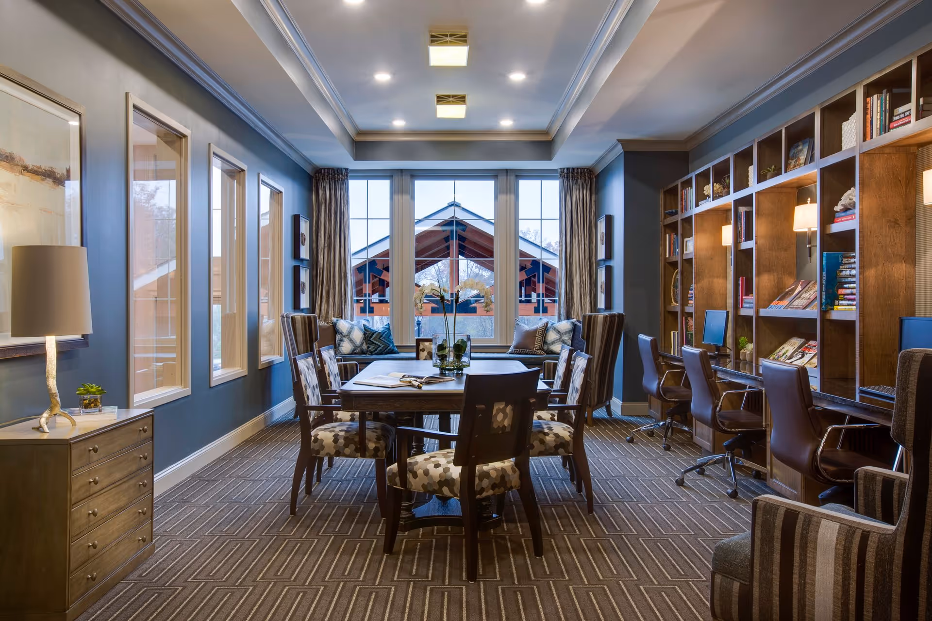 A well-lit interior room with a central wooden table surrounded by six patterned chairs. The room features large windows at the far end with a view of a building's roof outside. On the right side, there is a built-in wooden bookshelf with books, decorative items, and three computer workstations with chairs. On the left side, there are framed artworks on the wall and a small wooden cabinet with a lamp and a small plant on top. The room has a carpeted floor with a geometric pattern and recessed ceiling lights.