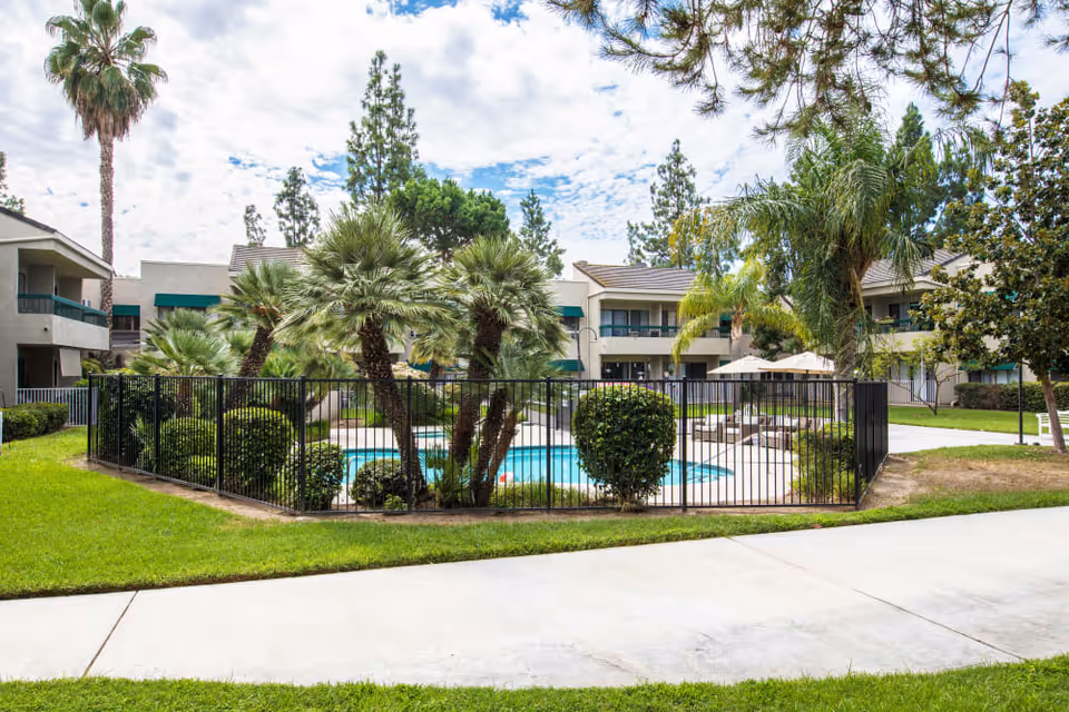 Outdoor view of a senior living facility named Citrus Place featuring a fenced swimming pool surrounded by palm trees and bushes, with two-story buildings in the background under a partly cloudy sky.