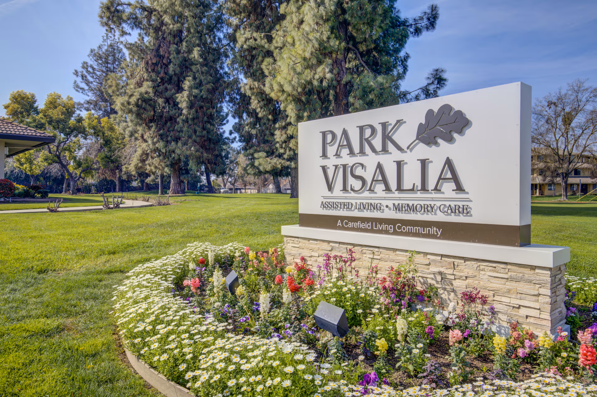 Outdoor view of a large sign for Park Visalia Assisted Living & Memory Care, surrounded by a flower bed with colorful flowers and green grass, with tall trees and a clear blue sky in the background.