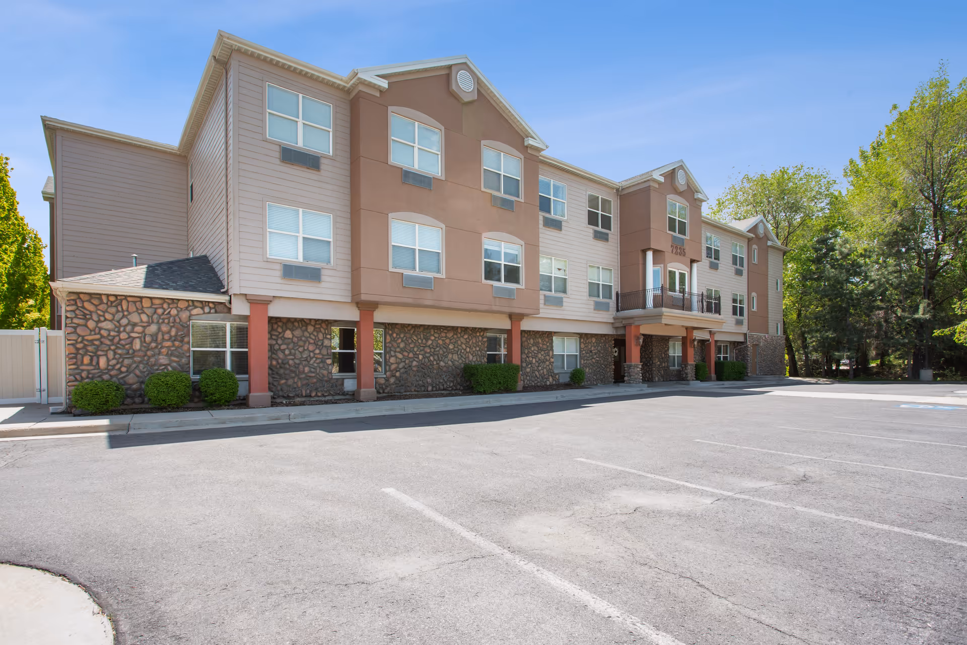 Exterior view of a three-story senior living facility building with a stone and beige facade, multiple windows, and a small balcony above the entrance. The building is surrounded by trees and has an empty parking lot in front under a clear blue sky.
