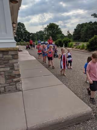 A group of young children walking outside along a paved path next to a building with stone and white siding. The children are accompanied by adults and are dressed in casual summer clothes. The background shows trees, bushes, and a cloudy sky.
