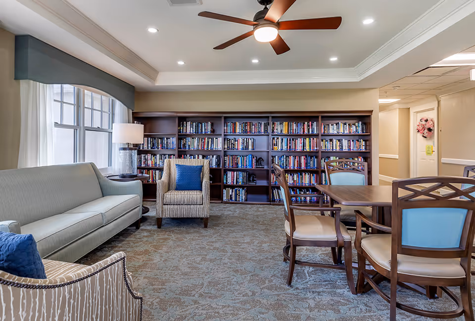 A cozy senior living facility common area with a large bookshelf filled with books against the back wall. The room features a light gray sofa, an armchair with a blue cushion, a wooden table with four chairs, and a ceiling fan with a light. There is a window with sheer white curtains and a blue valance on the left side, and a lamp on a side table next to the sofa.