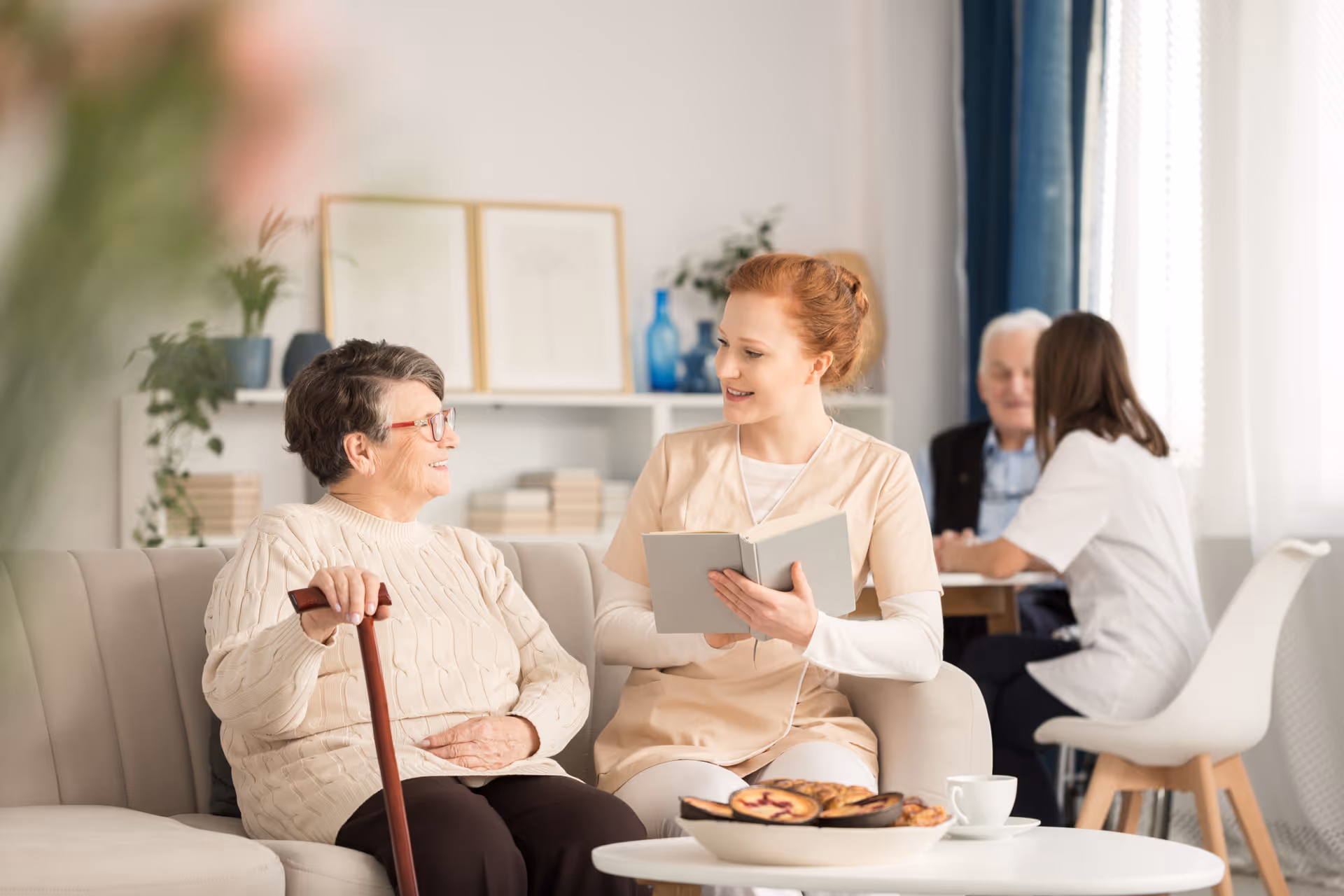 An elderly woman with a cane sitting on a couch and smiling while talking to a young female caregiver who is holding a book. In the background, another caregiver is interacting with an elderly man at a table in a bright, cozy room with plants and framed pictures on shelves.