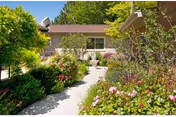 A garden pathway surrounded by lush greenery and colorful flowers leading to a single-story building with beige walls and a brown roof under a clear blue sky.
