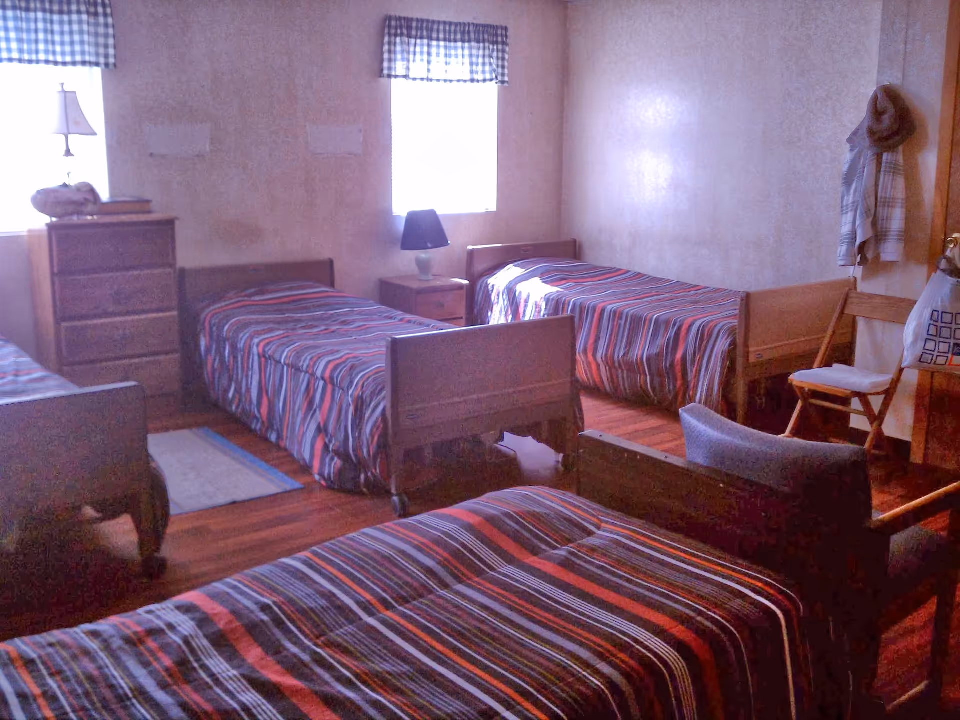 A bedroom with four single beds, each covered with striped bedspreads in shades of red, blue, and white. The room has wooden floors, two windows with checkered valances, a wooden dresser with a lamp on top, a nightstand with a lamp, a wooden chair, and a coat hanging on the wall.