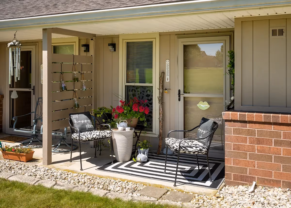 A small outdoor patio area with two black metal chairs featuring patterned cushions and pillows that say 'my happy place'. There is a small round table between the chairs with a teapot and cups. The patio has a black and white striped rug, potted plants with red flowers, and a wind chime hanging from the ceiling. The patio is attached to a beige building with a glass door and window, and a brick wall section is visible on the right.