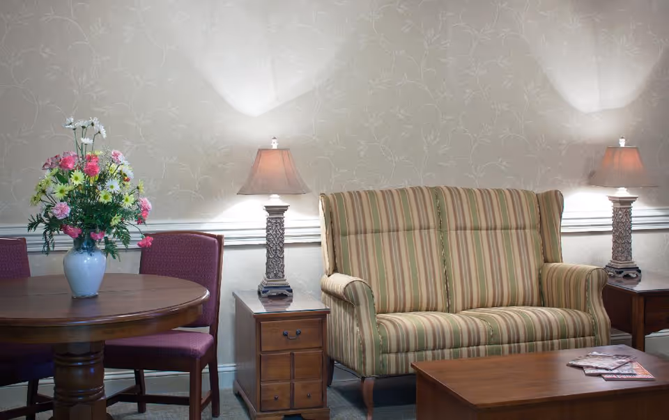 A cozy living room area with a striped upholstered loveseat, two wooden side tables each with a decorative lamp, a wooden coffee table with magazines, and a round wooden table with purple chairs and a vase of colorful flowers on top. The walls have a subtle floral patterned wallpaper.