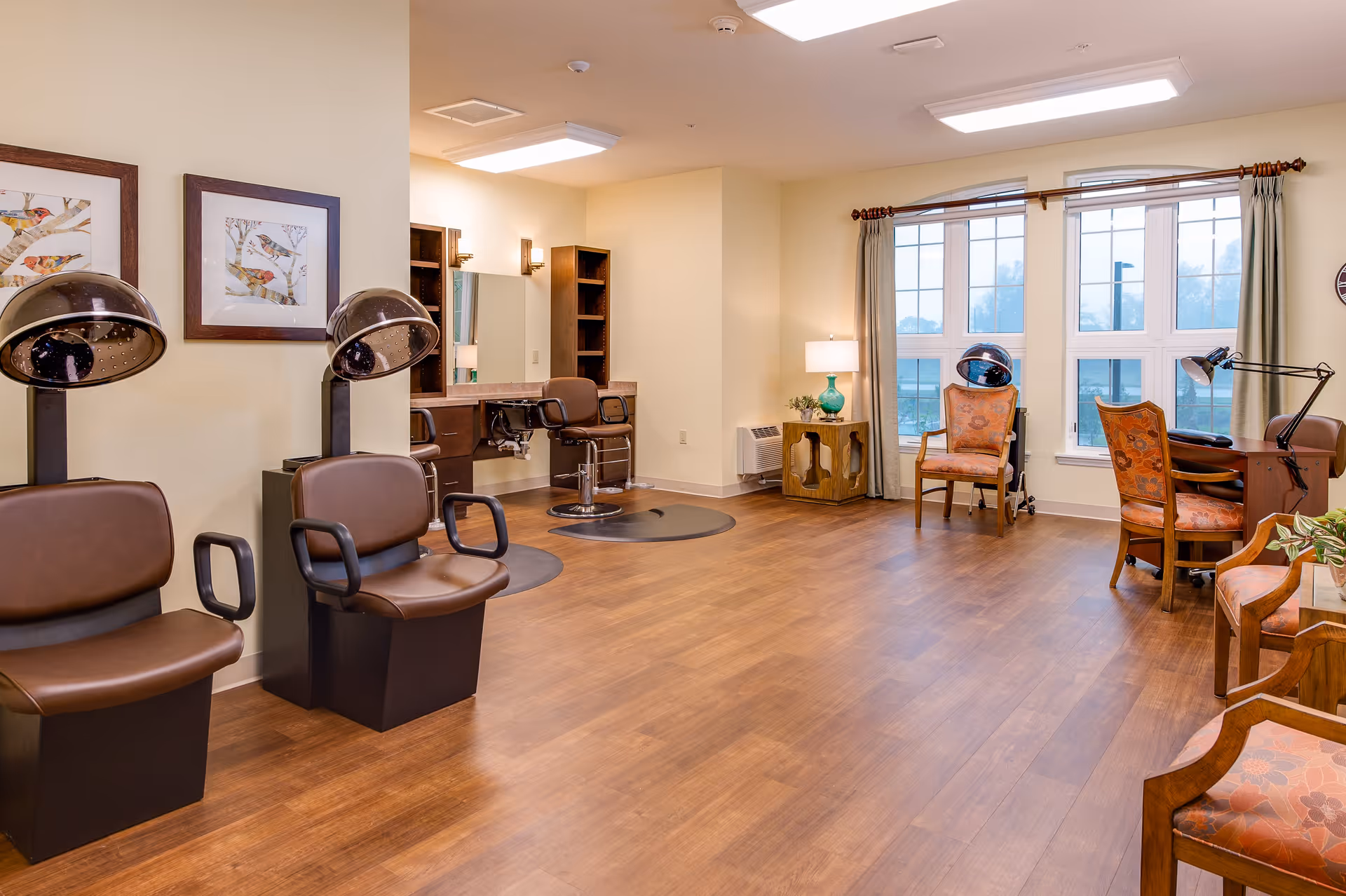 A spacious salon area with multiple brown leather salon chairs, hair drying stations, a vanity with mirrors and chairs, large windows with curtains, wooden flooring, and framed bird artwork on the wall.