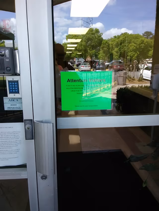 Glass entrance door of Crowne Health Care of Eufaula with a green sign for visitors and a keypad entry system. The door reflects the parking lot with several cars and trees under a partly cloudy sky.