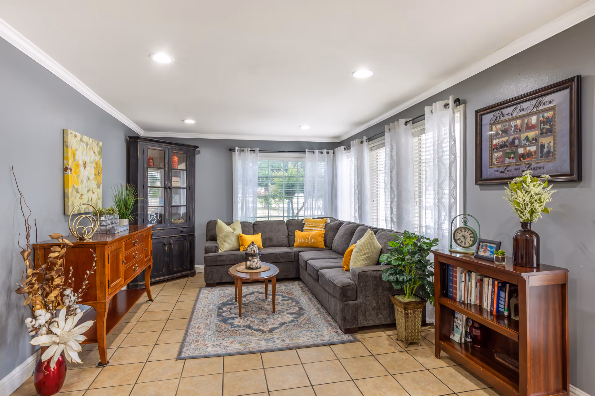 A cozy living room with a large gray sectional sofa adorned with yellow and beige pillows, a round wooden coffee table on a patterned rug, and multiple windows with sheer white curtains letting in natural light. The room features gray walls with white crown molding, a wooden sideboard with decorative items, a black corner cabinet, a wooden bookshelf with books and decor, and several green plants adding a touch of nature.