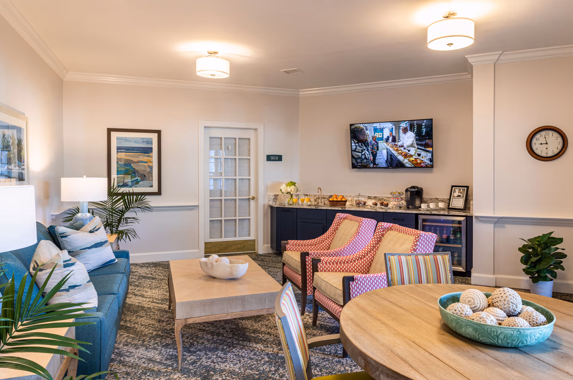 A cozy living room area in a senior living facility featuring a blue sofa with patterned pillows, two red patterned armchairs, a wooden coffee table with decorative items, a round wooden table with striped chairs, and a bowl with decorative balls. The room has a carpeted floor, framed artwork on the walls, a wall-mounted TV showing a cooking scene, a clock, plants, and a counter with refreshments and a coffee maker.