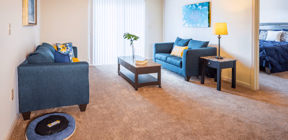 A cozy living room with two blue sofas facing each other, a wooden coffee table with a vase and book, a side table with a lamp, and a pet bed on the carpeted floor. A bedroom with a bed is visible through an open doorway.