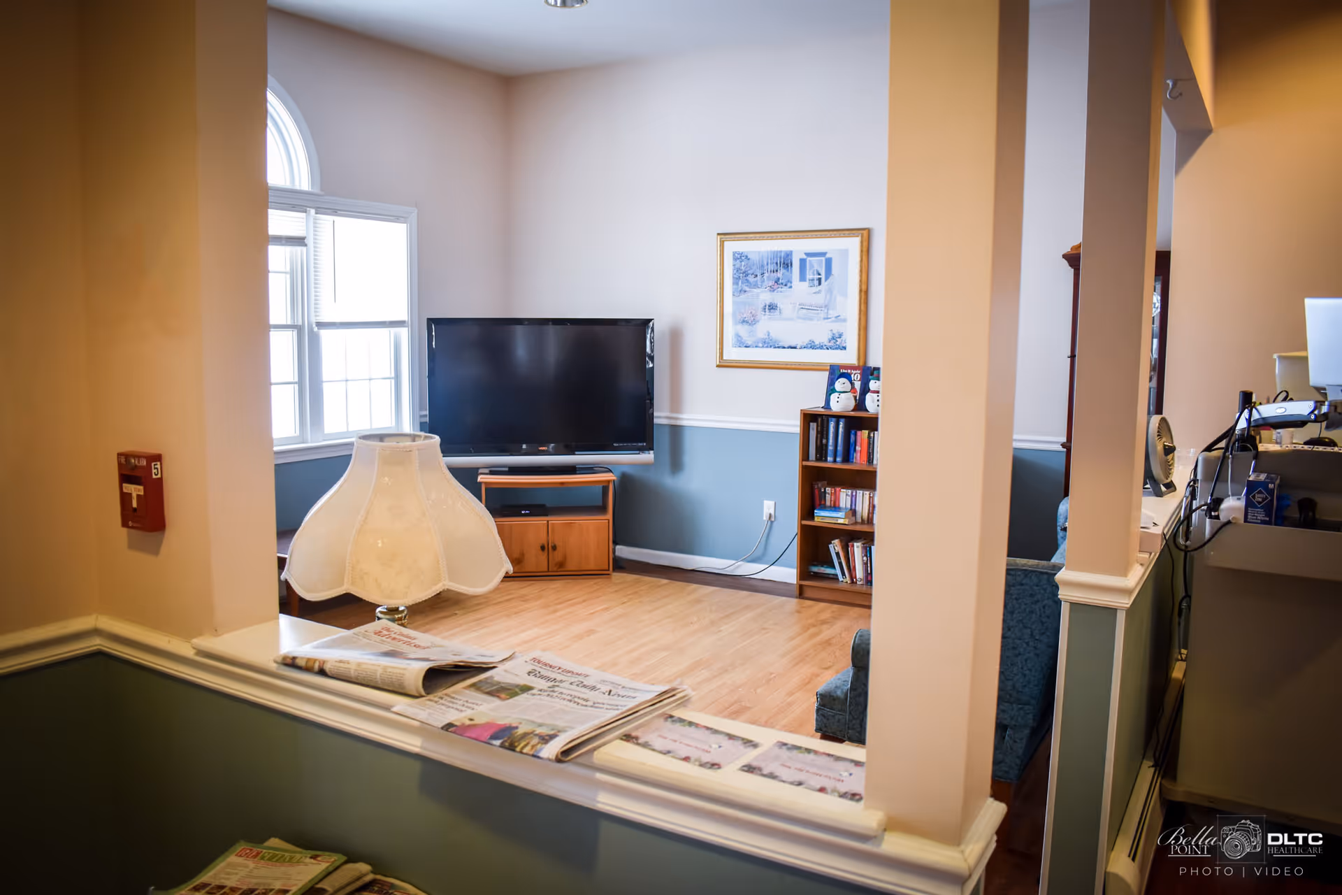 Interior view of a living room area with a flat-screen TV on a wooden stand, a bookshelf with books and decorative items, a large window letting in natural light, a table lamp with a white lampshade, and newspapers placed on a ledge in the foreground. The walls are painted beige and blue with white trim.