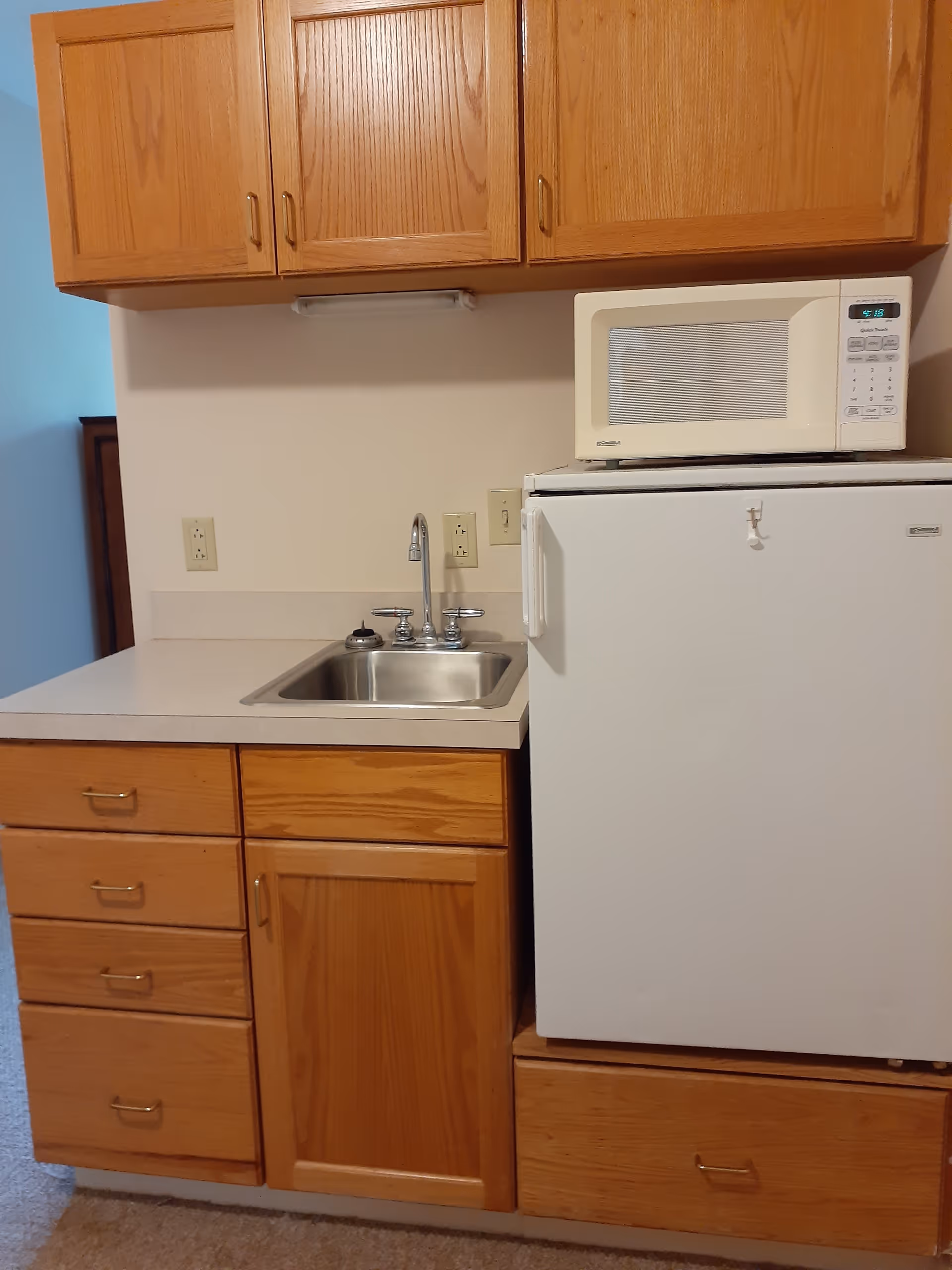 A small kitchenette area with wooden cabinets, a stainless steel sink with a faucet, a white mini refrigerator, and a white microwave placed on top of the refrigerator.