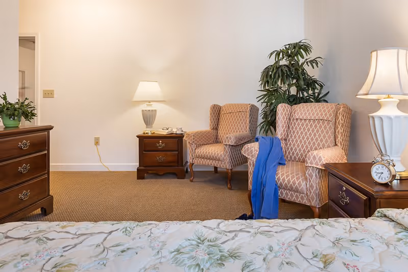 A bedroom sitting area with two patterned armchairs, wooden nightstands with lamps, a bed in the foreground and a potted plant.