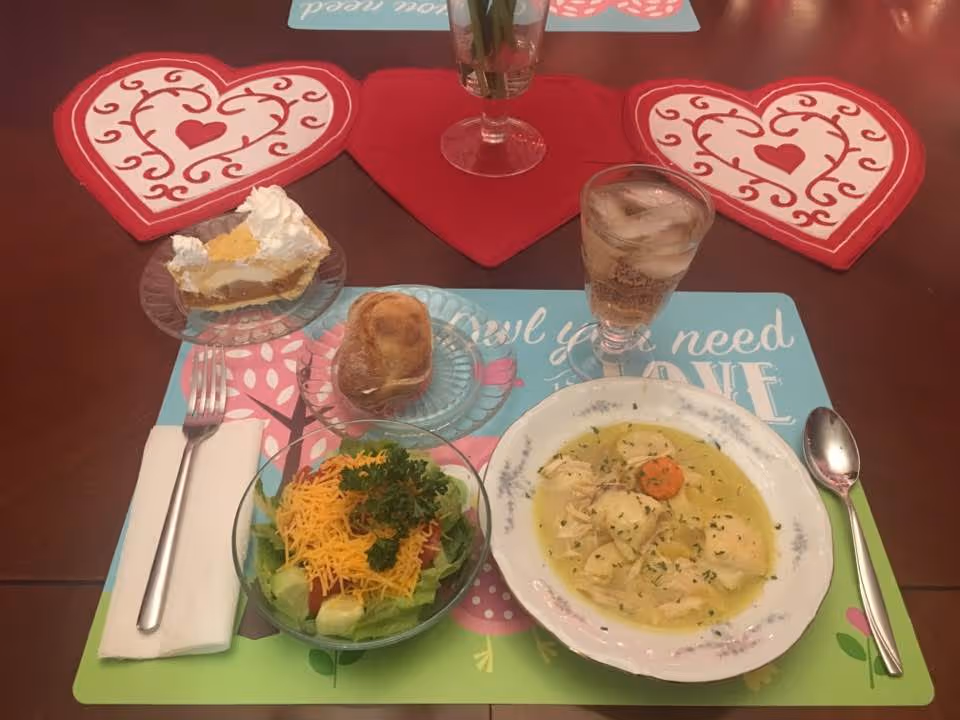 A dining table set with a meal including a bowl of chicken and dumplings soup, a glass of iced beverage, a small salad topped with shredded cheddar cheese and parsley, a dinner roll, and a slice of pie with whipped cream. The table has heart-shaped red placemats and a floral placemat with the words 'All you need is LOVE'. A fork and spoon are placed on either side of the placemat.