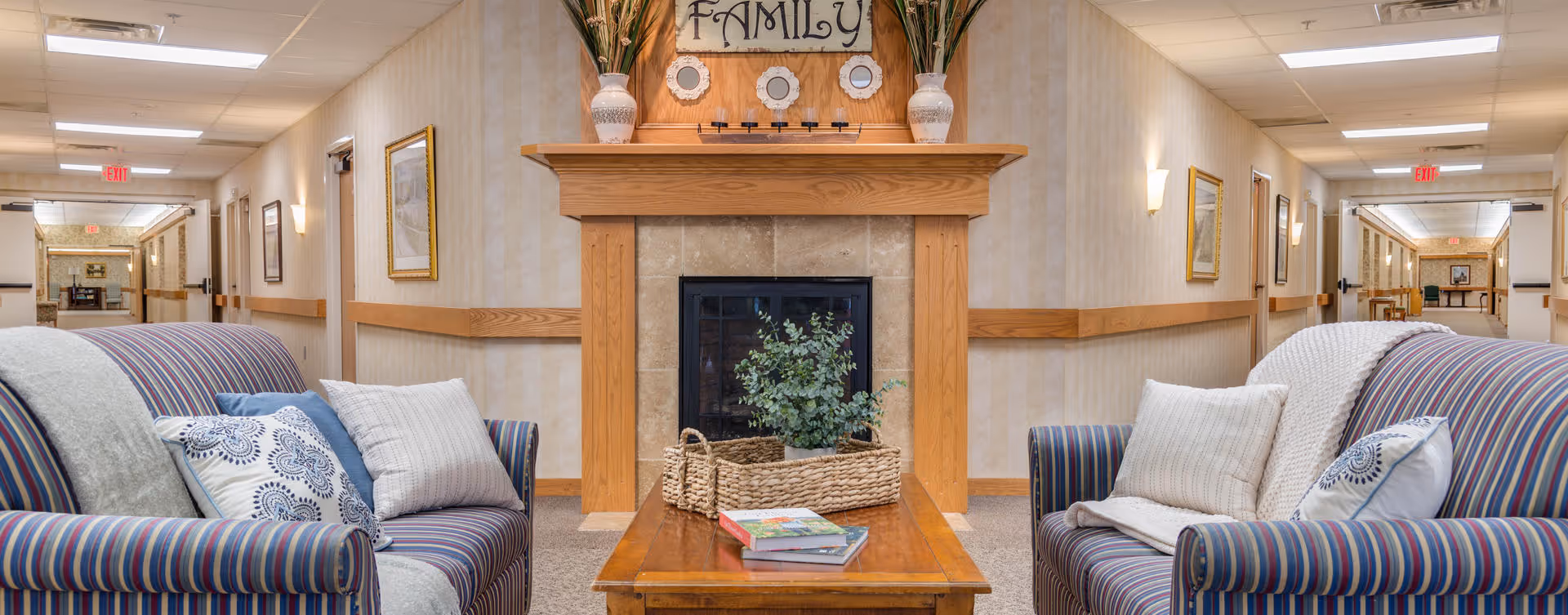 A cozy common area in a senior living facility featuring two striped sofas with cushions and throws facing each other across a wooden coffee table. The coffee table holds a woven basket with greenery and some books. Behind the table is a fireplace with a wooden mantel decorated with vases and a sign that reads 'FAMILY'. The space is well-lit with ceiling lights and wall sconces, and hallways extend on both sides of the room.