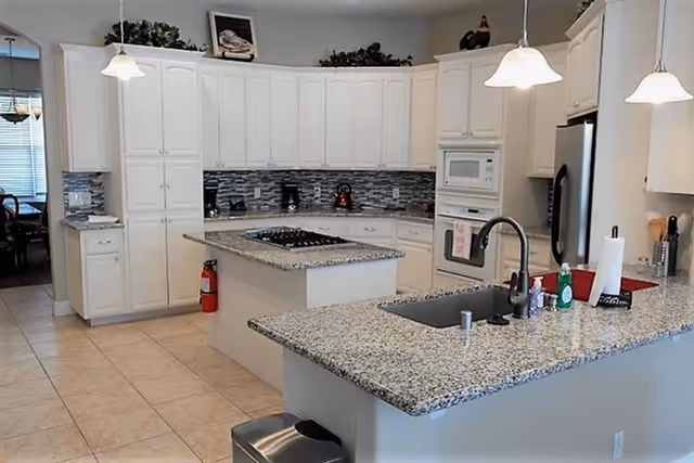 Bright kitchen with white cabinets, granite countertops, a central island with a gas stove, a sink with a modern faucet, and stainless steel appliances including a refrigerator and built-in microwave. Three pendant lights hang above the island, and there is a fire extinguisher mounted on the wall near the cabinets.