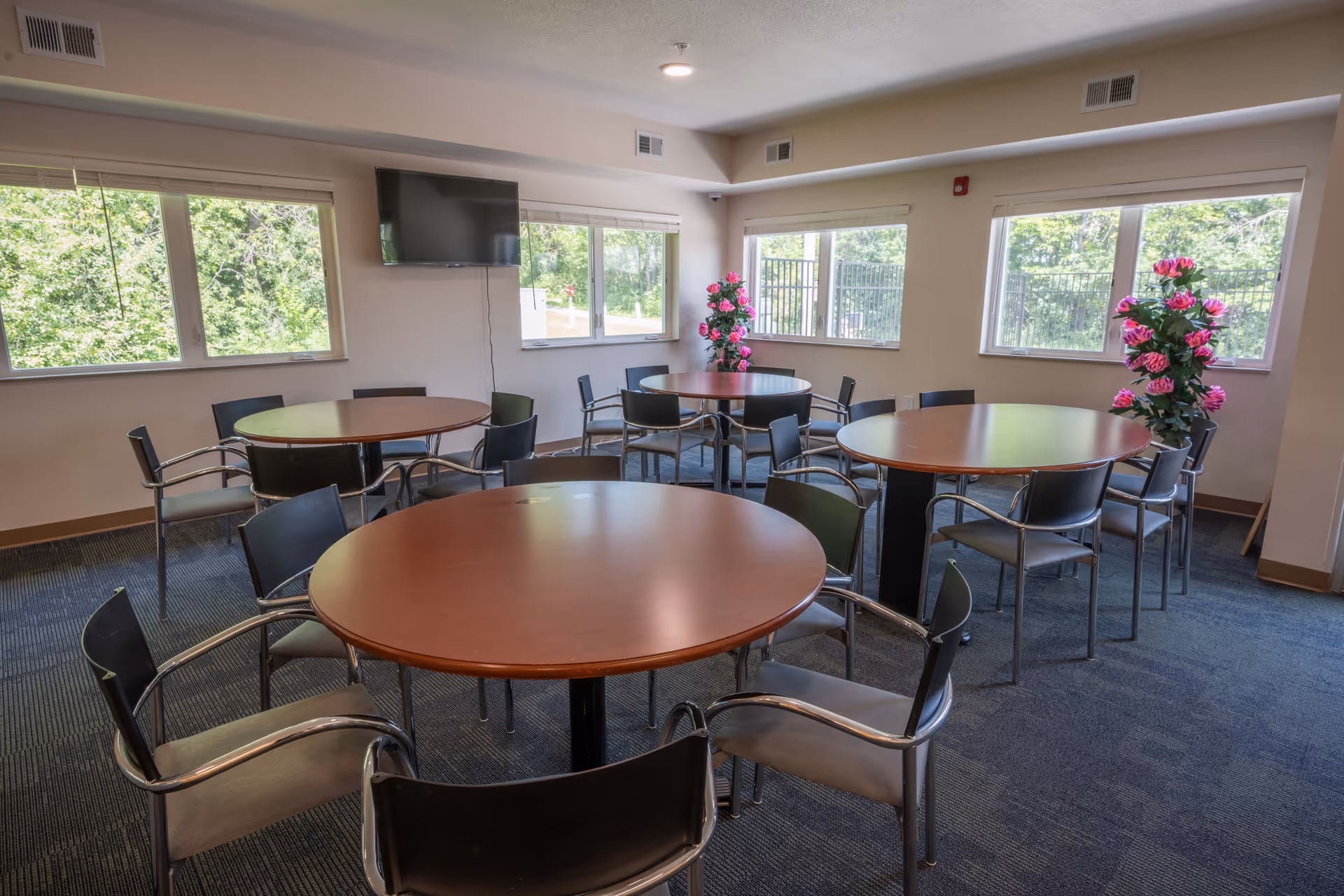 A bright common room with round wooden tables and metal chairs, large windows, a wall-mounted TV, and potted pink flowers.