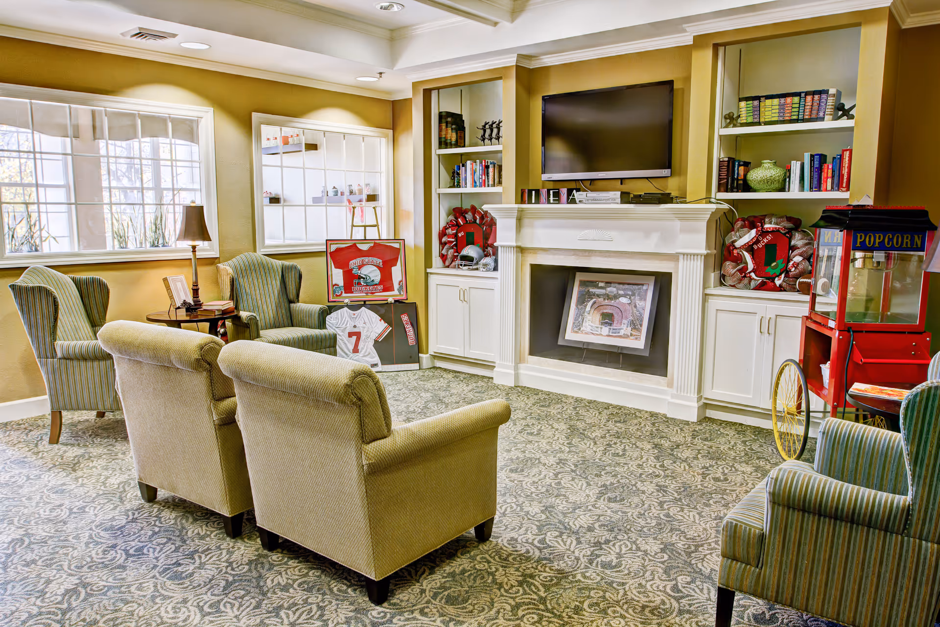A cozy living room area with four upholstered armchairs arranged around a small round table with a lamp and books. The room features a carpet with a floral pattern, built-in white shelves filled with books and decorative items, a wall-mounted flat-screen TV above a white fireplace, and framed sports memorabilia including jerseys and helmets. A red popcorn machine is visible on the right side near one of the armchairs. Large windows allow natural light to brighten the space.