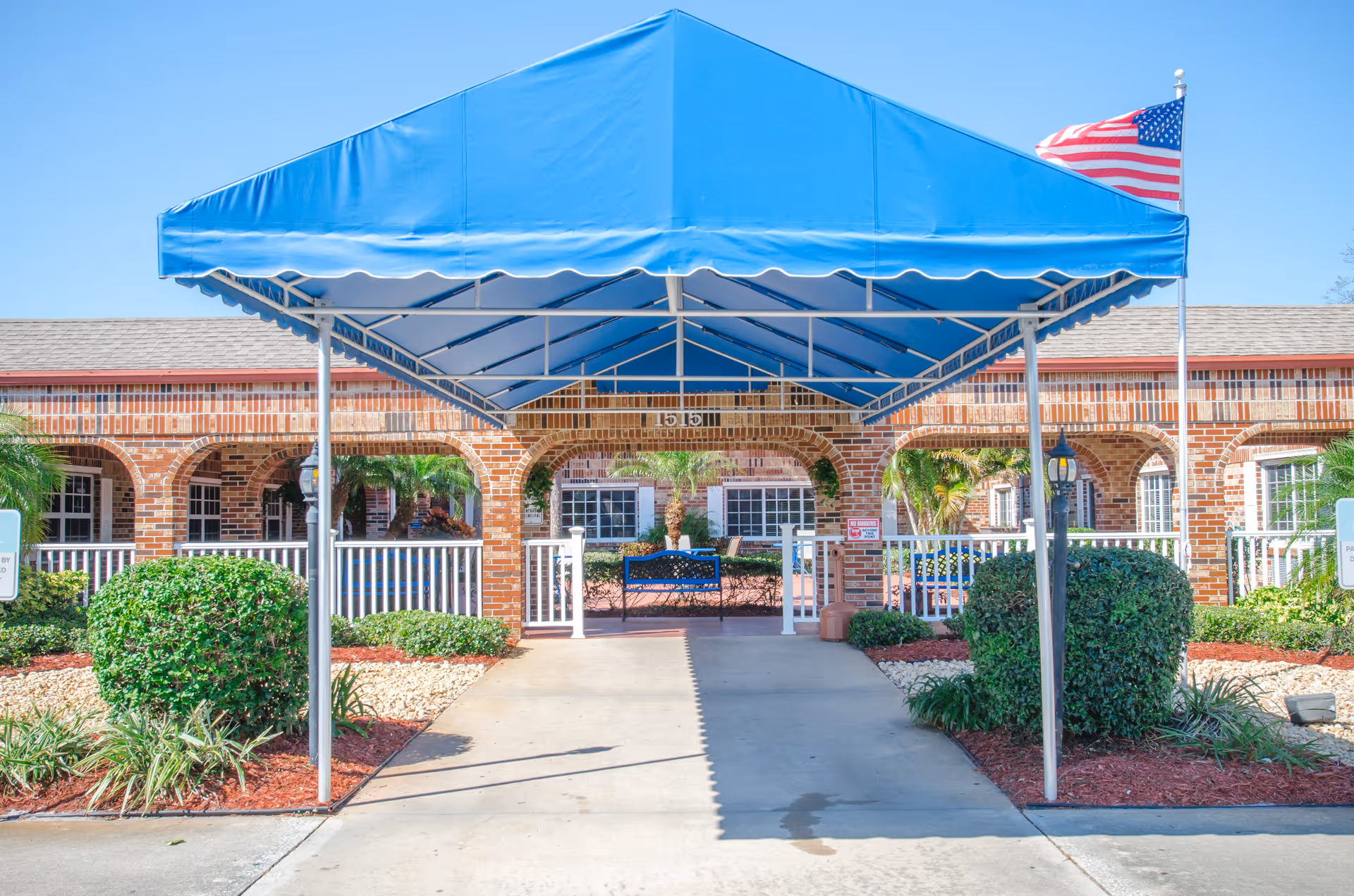 Entrance of a brick building with a blue canopy covering the walkway. There are neatly trimmed bushes and plants on either side of the path, a white fence, and an American flag on a flagpole to the right.