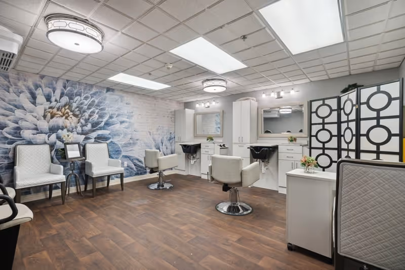 A bright and clean salon area in a senior living facility featuring two white salon chairs in front of sinks and mirrors, two white cushioned chairs with a small table between them, a decorative wall with a large floral mural, and a black and white room divider.