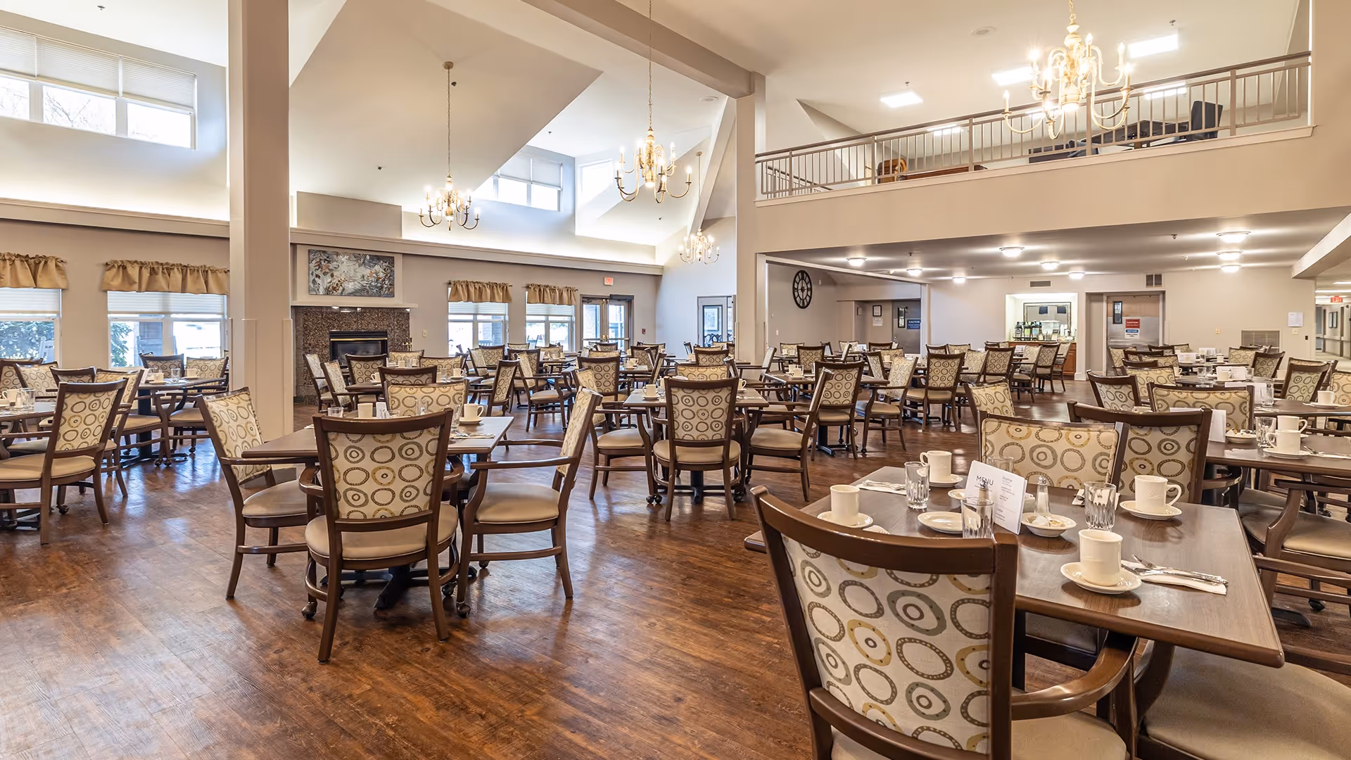 Spacious dining room with multiple wooden tables and chairs arranged neatly. The chairs have patterned upholstery with circular designs. The room features high ceilings with chandeliers, large windows with curtains, and a fireplace on one side. The floor is wooden, and the tables are set with cups, glasses, and utensils.