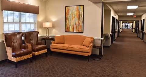 Carpeted interior hallway with a seating area containing two leather armchairs, an orange sofa, side tables, a lamp, and wall art.