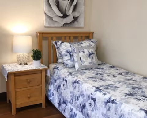 A single bed with a wooden headboard, covered with a floral patterned bedspread and matching pillows in shades of blue and white. Next to the bed is a wooden nightstand with two drawers, a white table lamp, and a small potted plant. Above the bed hangs a black and white picture of a rose. The room has light-colored walls and wooden flooring.