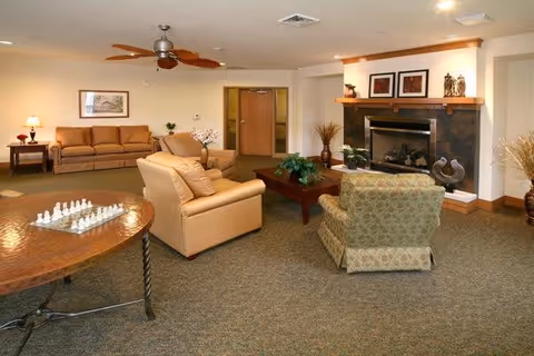 A cozy living room area with a beige sofa, a patterned armchair, a wooden coffee table with plants, and a fireplace with framed artwork above it. There is also a round table with a chess set in the foreground and a ceiling fan overhead.