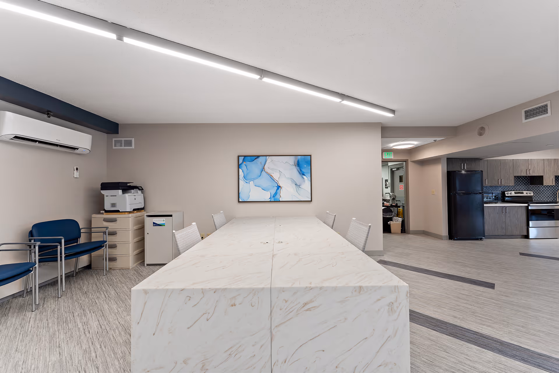 A modern communal room featuring a long marble table with white chairs around it. On the left side, there are blue chairs, a printer on a small cabinet, and an air conditioning unit mounted on the wall. In the background to the right, there is a kitchen area with dark cabinets, a black refrigerator, and stainless steel appliances. The walls are light beige, and a blue and white abstract painting hangs on the wall above the table.