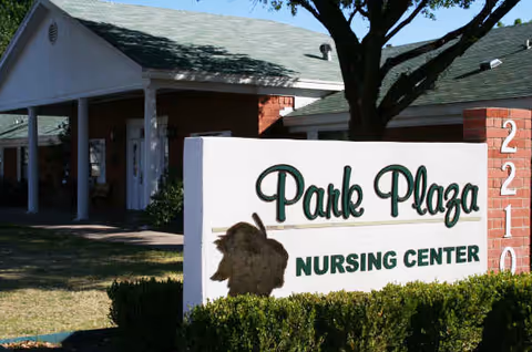 Exterior view of Park Plaza Nursing Center with a white sign displaying the facility name and address number 2210, a large tree, and part of the building with a covered entrance.
