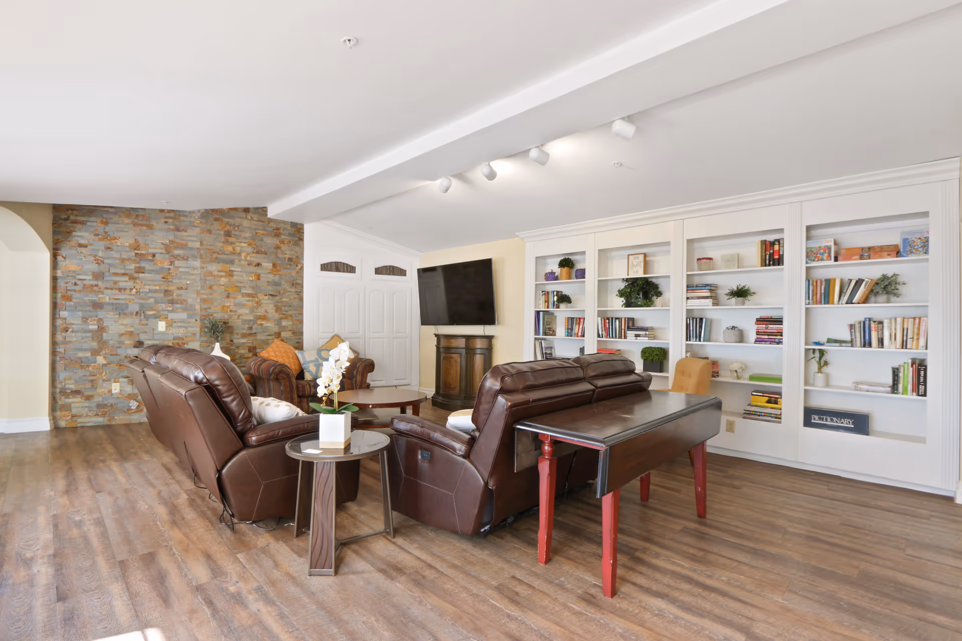 A cozy living room area with brown leather recliners, a round wooden coffee table, and a side table with a white orchid plant. The room features a stone accent wall, a mounted flat-screen TV above a small wooden cabinet, and built-in white bookshelves filled with books and decorative plants. The floor is wood, and the ceiling has recessed lighting.