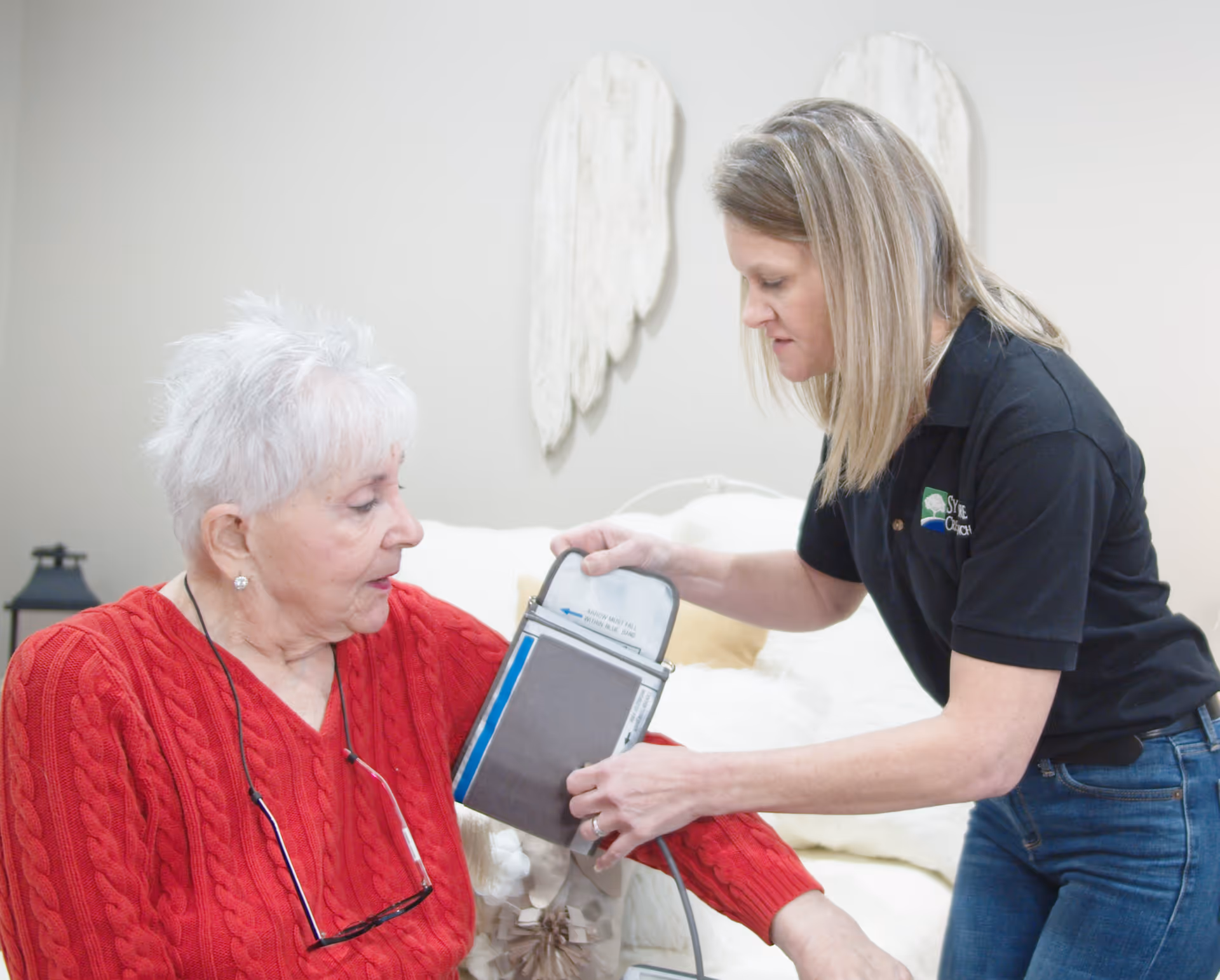 A caregiver in a black polo shirt is taking the blood pressure of an elderly woman wearing a red sweater in a cozy room with white walls and decorative angel wings on the wall behind them.